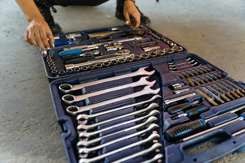 Close-up view of an open toolbox on a concrete floor, displaying various hand tools including wrenches, screwdrivers, and socket sets.